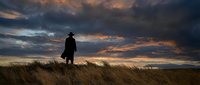 Silhouette of lone cowboy standing in golden grassland field under dramatic stormy sunset sky. Western frontier landscape with moody clouds and warm light.