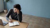 Young stressed businessman holding head in hands while working at desk with laptop and notebook in modern office interior.