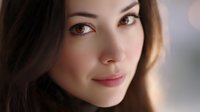 Young Asian woman with gentle smile and natural makeup looking at camera against blurred background, soft lighting highlights facial features and brown eyes.