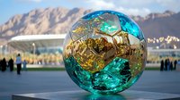 Metallic turquoise and gold sphere sculpture reflecting mountain landscape and people, modern art installation in public square against dramatic peaks.