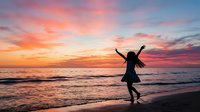 Silhouetted female figure with arms raised running along sandy beach at sunset, ocean waves in background, golden and pink sky, freedom and joy concept for lifestyle and travel content.