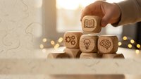 Hand placing wooden cube with book icon among blocks featuring lightbulb, gears and brain symbols representing education, innovation and learning concepts.