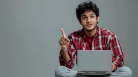 Young South Asian man in red plaid shirt sitting cross-legged with laptop, raising index finger in gesture of explanation or making a point against gray background.