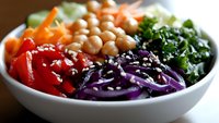 Fresh vegetable buddha bowl with chickpeas, kale, red cabbage, carrots, tomatoes and sesame seeds in white ceramic bowl, healthy plant based meal.