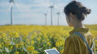 Young Asian woman in yellow jacket looking at wind turbines while standing in blooming rapeseed field, holding digital tablet, sustainable future concept.