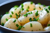 Steamed baby potatoes garnished with fresh chopped chives and black pepper in dark ceramic bowl, close-up view showing glossy surface and herb details.
