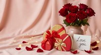 Romantic Valentine's Day scene with red roses bouquet, heart-shaped gift box with golden ribbon, love you card and scattered petals on pink silk backdrop.