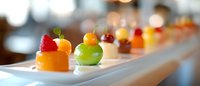 Colorful assortment of elegant mini desserts on white rectangular plate, featuring fruit-topped pastries with raspberry, orange, green apple, and cherry garnishes.