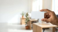 Male hand of African descent holding blank white business card in modern office environment with natural lighting and blurred background for corporate branding and professional networking purposes.