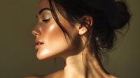 Young Asian woman with glowing skin and messy bun hairstyle in profile view against dark background, dramatic side lighting creates golden highlights on face.