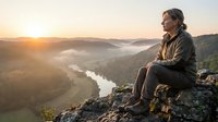 Middle-aged man with long dark hair and glasses sitting on rocky cliff edge overlooking misty river valley at sunrise, wearing brown jacket. Contemplative hiker enjoying scenic mountain landscape with forested hills and golden morning light.