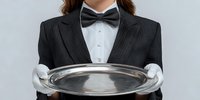 Professional server in formal attire with bow tie and white gloves holding empty silver serving tray against gray background.