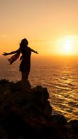 Woman with arms outstretched standing on rocky cliff overlooking golden sunset ocean, wearing flowing scarf, silhouetted against warm sky and water reflection for freedom and travel concepts.