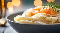 Fresh salmon and fettuccine pasta in creamy white sauce garnished with dill and black pepper, served in dark ceramic bowl against blurred lights.
