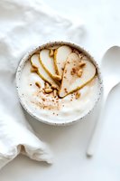 Creamy yogurt bowl topped with sliced pears, drizzled honey, pine nuts and cinnamon powder on white background, minimalist breakfast styling for healthy eating.