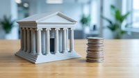 Miniature bank building model with classical columns next to stack of coins on wooden desk in modern office environment for financial concepts.