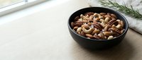 Mixed nuts and dried fruits in black bowl on wooden table with rosemary sprig for healthy snacking and cooking ingredients.