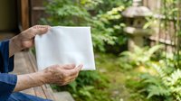 Senior man with weathered hands holding blank white paper outdoors against green foliage background, suitable for mockups, advertising, and promotional campaigns.