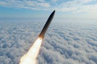 Missile launching through cloud layer with bright rocket engine flame trail against blue sky background, dramatic military defense concept.