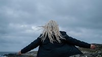 Young blonde woman with flowing hair standing with arms outstretched against dramatic cloudy sky, expressing freedom and joy in outdoor natural setting.