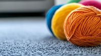 Colorful yarn balls in orange, yellow, blue and pink arranged on gray textured fabric background with selective focus on orange wool skein for knitting projects.