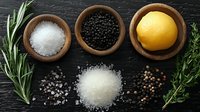 Fresh cooking ingredients arrangement with lemon, sea salt, black peppercorns in wooden bowls, scattered spices and aromatic rosemary sprigs on dark wooden surface.