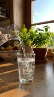 Water pouring into glass on wooden table by sunny window with potted plants and fruit bowl, suitable for beverage, wellness, and lifestyle content.