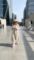 Woman with reddish-brown hair wearing cream trench coat and straw hat standing in modern urban plaza surrounded by contemporary glass and concrete buildings on sunny day.