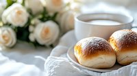 Freshly baked sweet buns with powdered sugar served with coffee and white roses in background, creating elegant breakfast atmosphere.