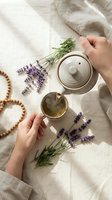 Hands pouring herbal lavender tea from ceramic teapot with fresh purple flowers and wooden beads on linen fabric for relaxation and wellness rituals.