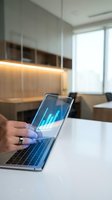 Person holding tablet displaying blue financial data charts in modern office workspace with warm lighting and contemporary furniture.