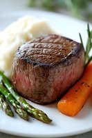 Juicy grilled beef tenderloin steak served with mashed potatoes, fresh asparagus, and glazed carrots on white plate, garnished with rosemary sprig.