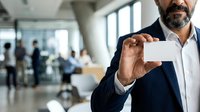 Mature male professional with dark beard wearing navy blazer and white shirt holding blank business card in modern office lobby with colleagues in background.