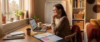 Woman with long dark hair and light skin tone, wearing glasses and casual beige sweater, working at wooden desk with laptop, notebook, and coffee cup in warm, sunlit home office with shelving, plants, and cozy decor.