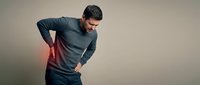 Young Caucasian man in gray sweater experiencing lower back pain, holding his spine with red inflammation highlight indicating discomfort and injury.