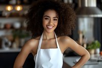 Young African American woman barista with natural curly afro hair wearing white apron smiling confidently in modern coffee shop interior with warm lighting.