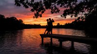 Romantic couple silhouette embracing on wooden dock at sunset over calm lake with dramatic orange and purple sky creating peaceful evening atmosphere.