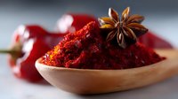 Red chili powder in wooden spoon with star anise and dried red chilies in background, macro shot showing vibrant texture and spicy seasoning detail.