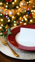 Festive holiday table setting with red charger plate, white napkin, gold cutlery, holly decoration, and blurred golden bokeh lights in background. Perfect for holiday dining, event planning, and seasonal marketing materials.