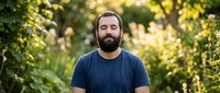Bearded man with dark hair and facial hair wearing navy blue t-shirt outdoors surrounded by green foliage and natural sunlight in garden setting.