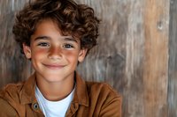 Cheerful Hispanic boy with curly hair wearing brown jacket and white t-shirt smiling warmly at camera against rustic wooden background.