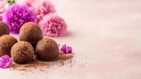 Dark chocolate truffles dusted with cocoa powder arranged with pink carnations and purple flower petals on pink textured background, selective focus for dessert menu.