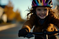Young girl with red hair wearing safety helmet smiles while riding bicycle during golden hour sunset. Active healthy lifestyle and childhood outdoor activities.