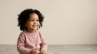 Toddler with curly dark hair and medium skin tone wearing pink sweater, smiling while looking upward against neutral background for childcare and family content.