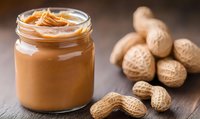 Creamy peanut butter in glass jar with whole peanuts scattered on rustic wooden surface, natural spread showing smooth texture and rich color.
