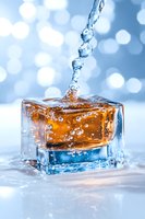 Crystal clear water drops falling into amber liquid in glass cube container creating splash effect against soft blue bokeh background with dynamic motion capture.