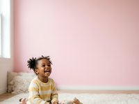 Happy toddler boy with dark curly hair, African descent, wearing yellow striped shirt, sitting on carpet in bright room with pink wall and natural light from window.