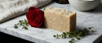 Handmade natural soap bar with oatmeal exfoliant displayed on marble with fresh red rose and thyme herbs for spa and wellness product marketing.