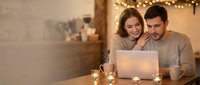 Young couple using laptop together at home with warm fairy lights creating cozy romantic atmosphere for evening browsing or online shopping.