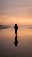 Woman in dark clothing walking alone on wet beach at sunset, silhouetted against golden sky and calm water. Solitude and contemplation concept for wellness and lifestyle use.